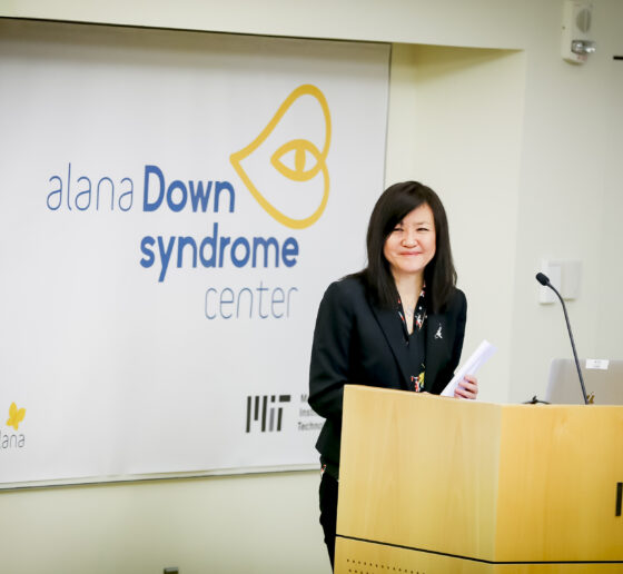 Li-Huei Tsai stands at an MIT podium with the Alana Center logo in the background