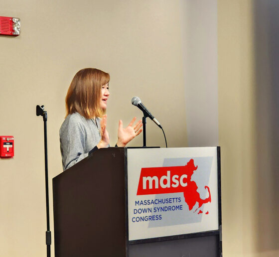 Li-Huei Tsai smiles and gestures from behind a podium that displays the Massachusetts Down Syndrome Congress logo