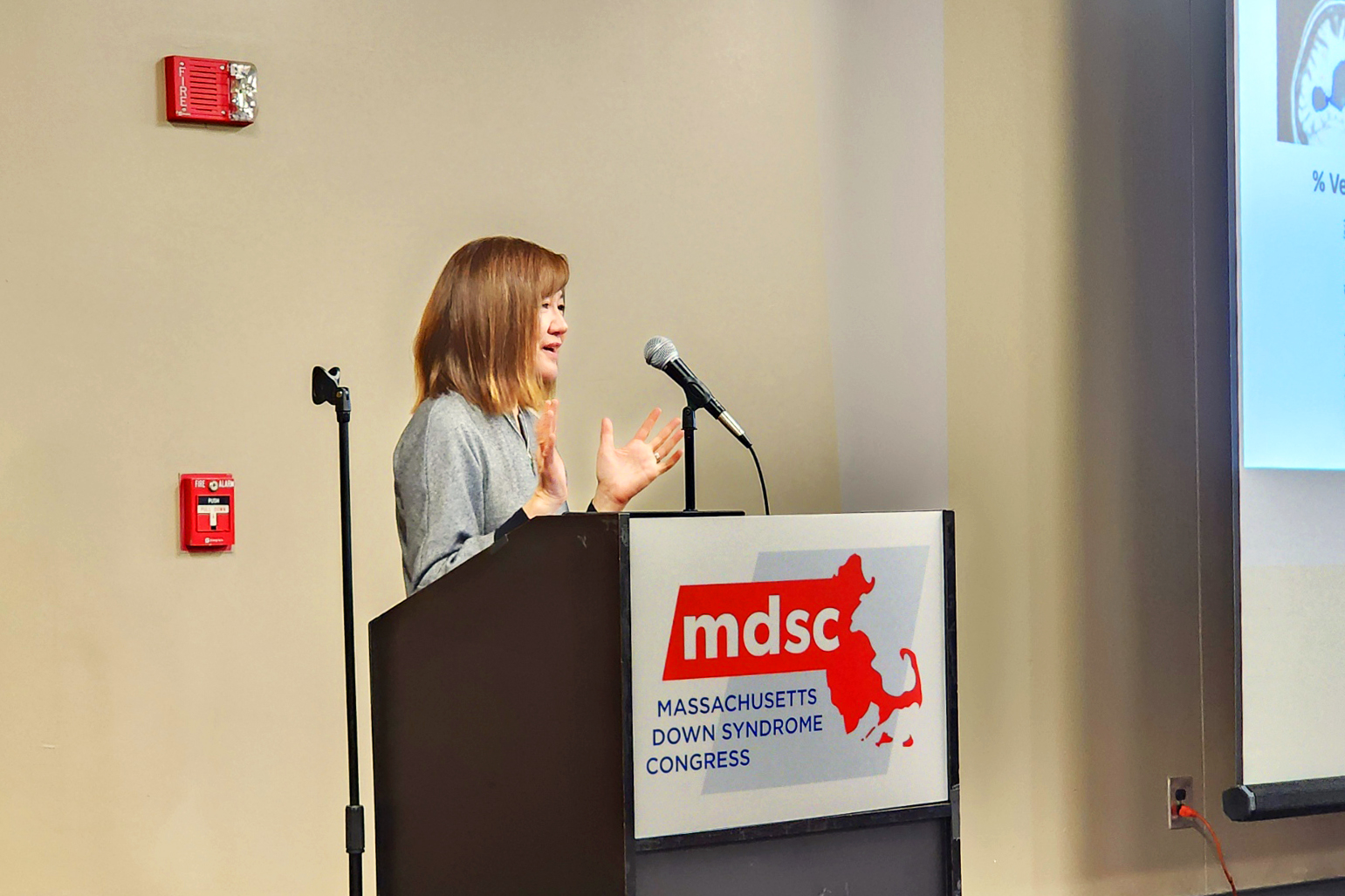 Li-Huei Tsai smiles and gestures from behind a podium that displays the Massachusetts Down Syndrome Congress logo